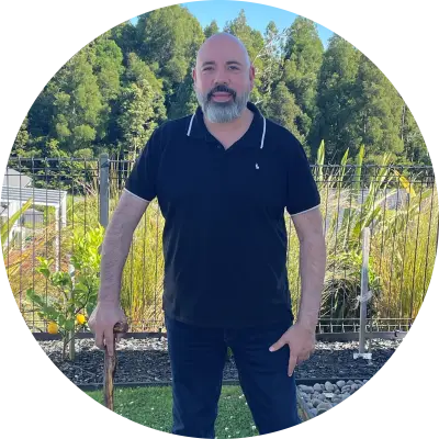 Geno Sisneros is smiling at the camera. He is a bald white man with a beard, wearing a black polo shirt and black trousers. He carries a wooden walking stick. .