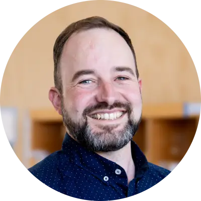 Karl Archibald is smiling. He has a beard and brown hair. He is wearing a blue shirt..