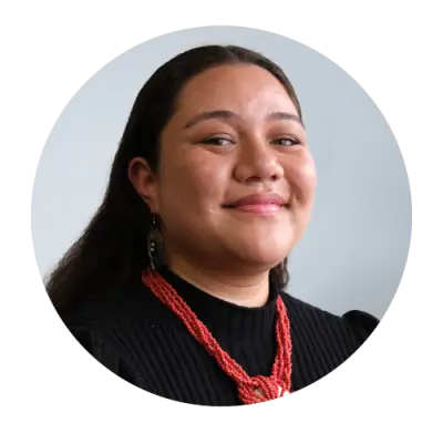 Nele Kalolo looks at the camera and smiles. She wears her hair loose and straight behind her ears. She wears a red necklace over a black top. .