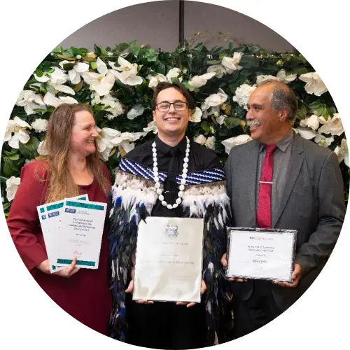 Jesse stands holding his graduation certificate, he is grinning from ear to ear and holding his graduation certificate. Jesse wears a Māori cloak and is flanked by two older people, also smiling and holding certificates.. .