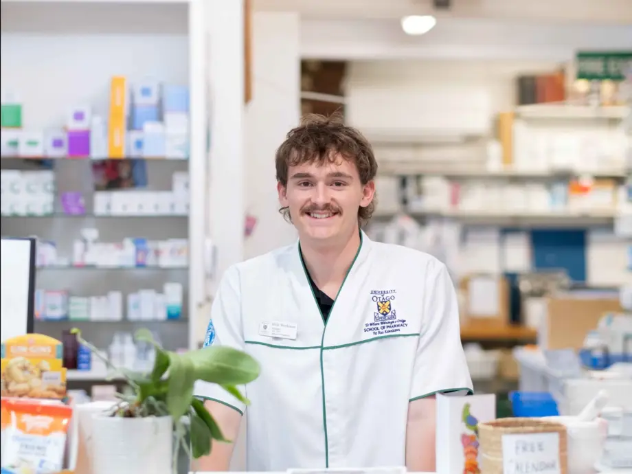 Nick is in a Pharmacy; you can see medicines behind him. He is wearing a white Pharmacist shirt and has a fantastic moustache. He is smiling. .
