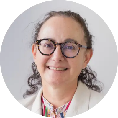 Nicola looks at the camera and smiles. She is wearing stylish tortoiseshell pattern rimmed glasses and has her curly brown hair swept back in a ponytail..