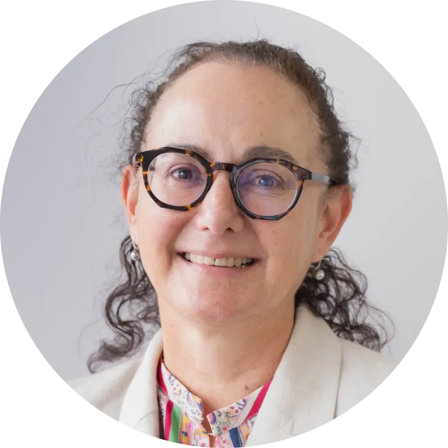 Nicola looks at the camera and smiles. She is wearing stylish tortoiseshell pattern rimmed glasses and has her curly brown hair swept back in a ponytail..