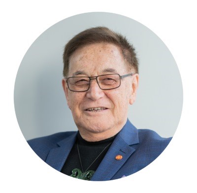 Pāpa Bill is smiling. He is wearing a black t-shirt, with a blue blazer, and has glasses on.. 