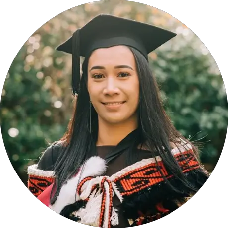 Sade is a young Māori wahine. She is facing the left, wearing a graduation cap and gown, with a korowai over the top. The korowai has koru detailing around the top, and is primarily black, red and white.  . 