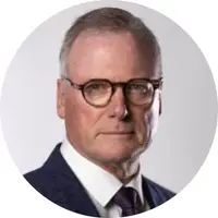A serious looking older white man. He wears glasses and smart suit with a dark tie..